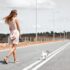 woman in white sleeveless dress holding harness of puppy walking on asphalt road during daytime