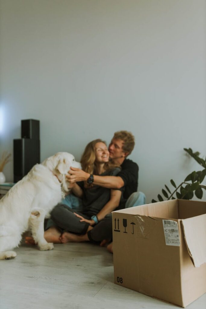 Anastasia Shuraeva - NeposlušnéTlapky Young couple sitting with a white dog during moving. A cardboard box is in the foreground.