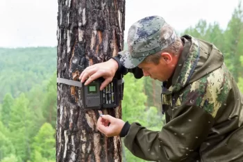 Muž v maskáčovém oblečení upevňuje fotopast na kmen stromu v lesní krajině