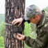 Muž v maskáčovém oblečení upevňuje fotopast na kmen stromu v lesní krajině